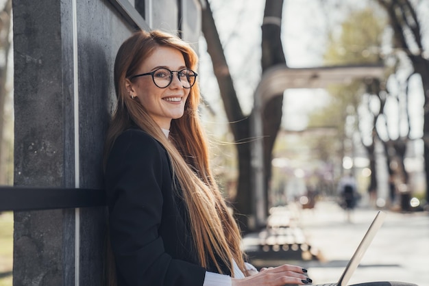 Mulher ruiva sentada no parque e posando enquanto trabalhava em seu projeto online conceito de trabalho online mulher trabalhando ao ar livre na natureza escritório móvel