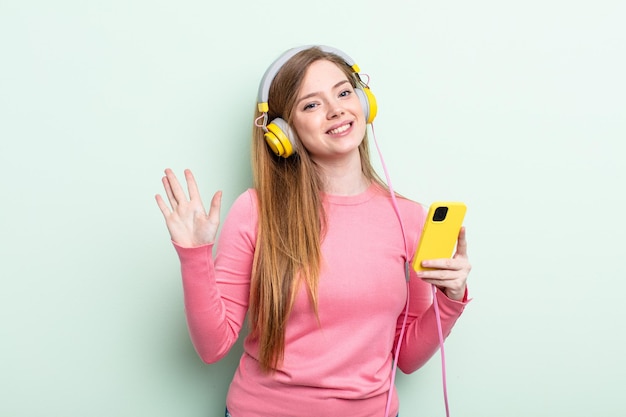 Mulher ruiva sorrindo alegremente, acenando com a mão, dando as boas-vindas e cumprimentando você. fones de ouvido e conceito de smartphone