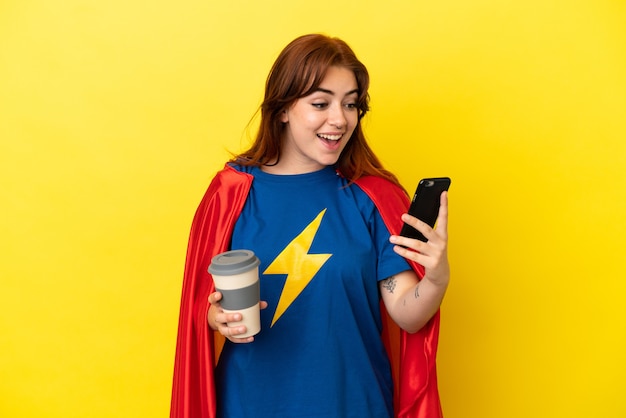 Mulher ruiva super-heroína isolada em um fundo amarelo segurando um café para levar e um celular