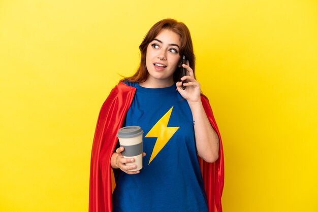 Mulher ruiva super-heroína isolada em um fundo amarelo segurando um café para levar e um celular