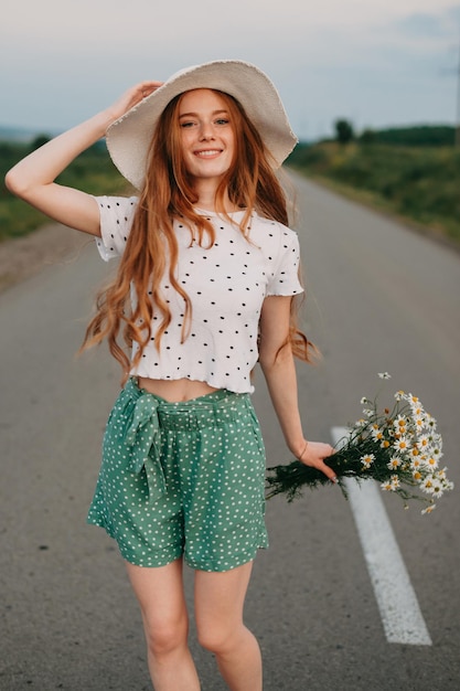 Foto mulher ruiva tendo um encontro fora da cidade segurando buquê de flores brancas e sorrindo usando chapéu de sol verão natureza vida no campo dia ensolarado