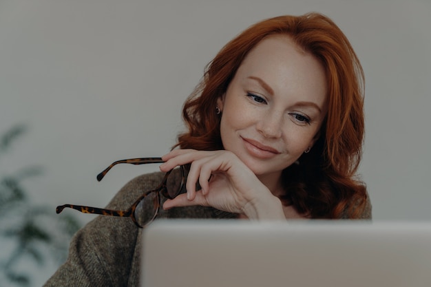 Mulher ruiva tira óculos, focada no monitor do computador portátil