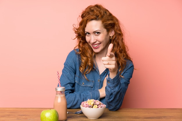 Mulher ruiva tomando café da manhã cereais e frutas aponta o dedo para você