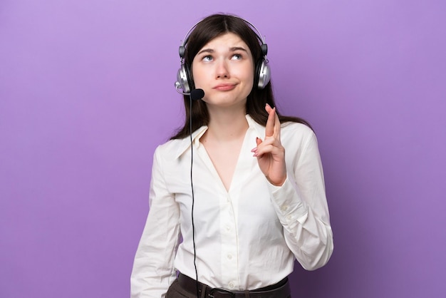 Mulher russa de telemarketing trabalhando com um fone de ouvido isolado em fundo roxo com dedos cruzando e desejando o melhor