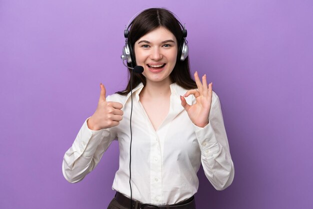 Mulher russa de telemarketing trabalhando com um fone de ouvido isolado em fundo roxo mostrando sinal de ok e polegar para cima gesto