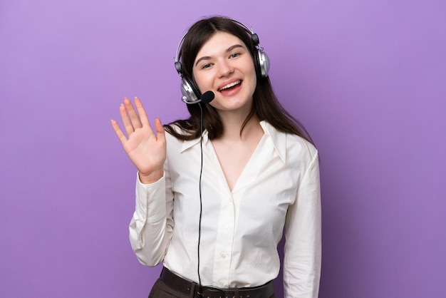 Mulher russa de telemarketing trabalhando com um fone de ouvido isolado em fundo roxo saudando com a mão com expressão feliz