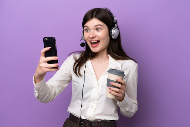 Mulher russa de telemarketing trabalhando com um fone de ouvido isolado em fundo roxo segurando café para levar e um celular