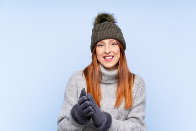 Mulher russa jovem ruiva com chapéu de inverno sobre aplaudindo a parede azul isolada