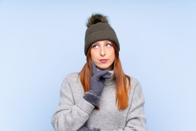 Mulher russa jovem ruiva com chapéu de inverno sobre fundo azul isolado, pensando uma idéia