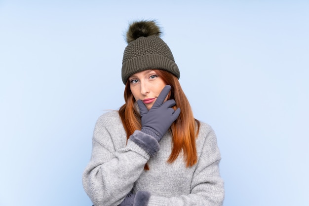 Mulher russa jovem ruiva com chapéu de inverno sobre parede azul isolada, pensando uma idéia