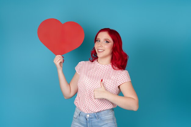 mulher satisfeita com cabelo vermelho segurando um grande coração de papel vermelho e fazendo sinal de positivo