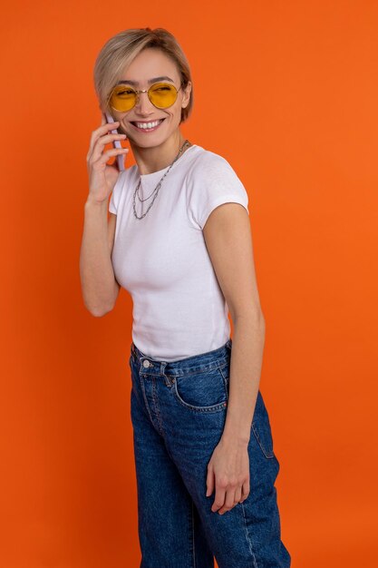 Foto mulher satisfeita com camiseta branca e calças jeans falando em telefone móvel isolada sobre fundo laranja