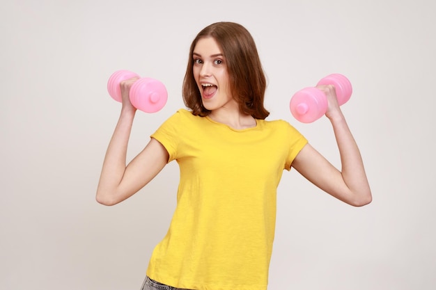 Mulher satisfeita e feliz em camiseta casual amarela fazendo exercícios esportivos com halteres rosa, bombeando músculos, expressando emoção. Tiro de estúdio interior isolado em fundo cinza.