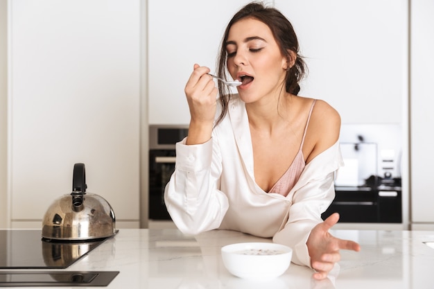 mulher saudável vestindo roupas de seda comendo granola com leite, enquanto toma café da manhã no apartamento