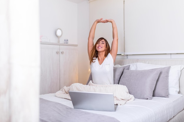 Mulher se alongando na cama depois de acordar de manhã pronta para trabalhar em casa. retrato de uma jovem sorridente, sentada com o laptop na cama. freelance trabalhando em casa conceito