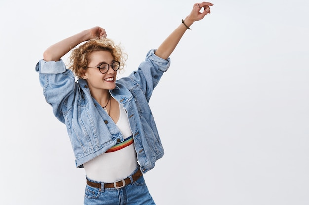 mulher se empolga com vibrações musicais dançando despreocupada sorrindo amplamente acenando e levantando as mãos de alegria feche os olhos e relaxe durante a festa incrível na pista de dança virando à direita vestindo jeans sobre a parede branca