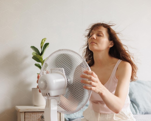 Mulher se refresca em casa com a ajuda de um ventilador de ar condicionado sentada na cama em frente a um elétrico