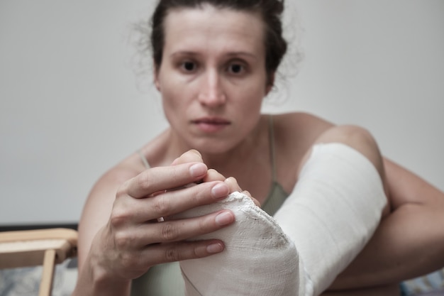 Foto mulher se senta na cama e segura uma perna quebrada. a perna está engessada. reabilitação domiciliar.