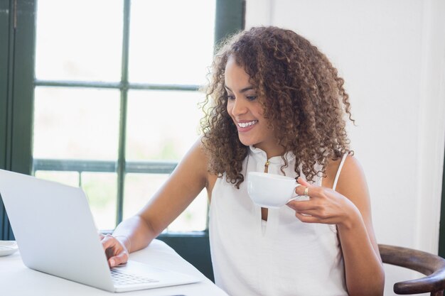 Mulher segurando a xícara de café e usando o laptop em um restaurante