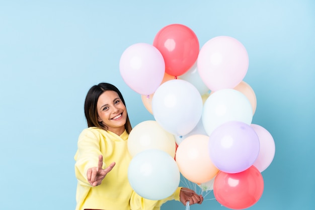 Mulher segurando balões em uma festa na parede azul sorrindo e mostrando sinal de vitória