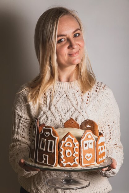 Mulher segurando bolo caseiro com fachadas de casa urbana de pão de gengibre, uma árvore de Natal e uma rena em um suporte de vidro