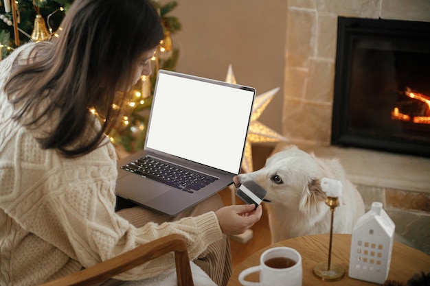 Mulher segurando cartão de crédito e laptop com tela em branco no fundo de um cachorro curioso fofo e sala de natal decorada festivamente Vendas de Natal e compras on-line Espaço para texto