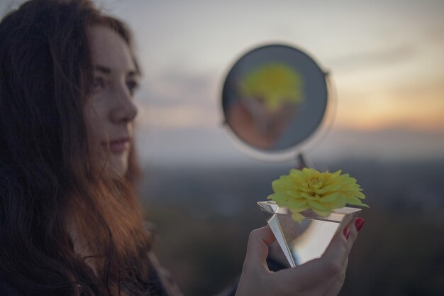 Foto mulher segurando espelho de mão com reflexo de cristal e flor durante o pôr do sol