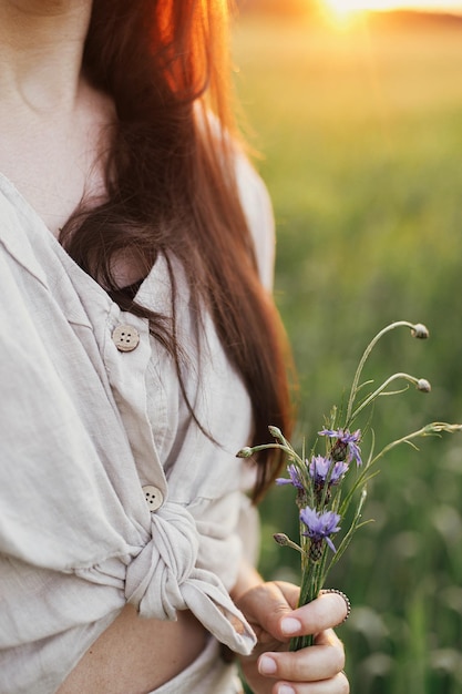 Mulher segurando flores silvestres no campo de trigo na luz quente do pôr do sol Fechar a jovem fêmea em vestido rústico com flores nas mãos no campo de verão à noite Momento atmosférico tranquilo