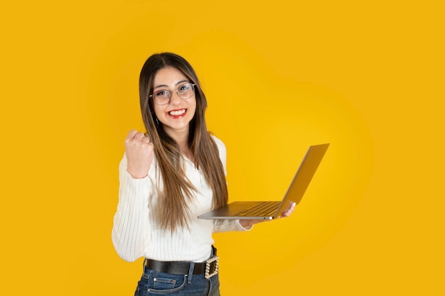 Mulher segurando laptop comemorando com gesto de vencedor em pé sobre fundo amarelo Copiar espaço