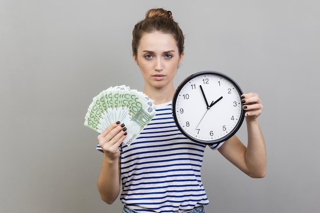 Mulher segurando o relógio de parede e diversão de notas de euro olhando para a câmera