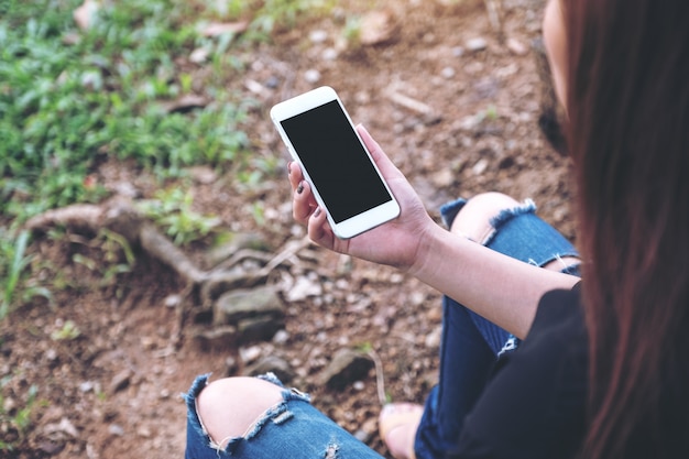 mulher segurando o telefone móvel branco com tela preta em branco, enquanto está sentado no chão ao ar livre
