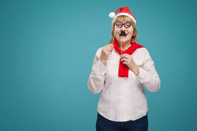 Mulher segurando óculos e lábios feitos de papel Uma mulher de meia-idade no ano novo e roupas de Natal em um fundo azul