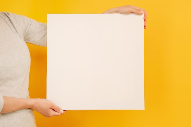 Mulher segurando papel de lona branca em branco com copyspace nas mãos na folha vazia de fundo amarelo ...