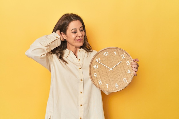 Foto mulher segurando relógio em pano de fundo amarelo tocando a parte de trás da cabeça pensando e fazendo uma escolha