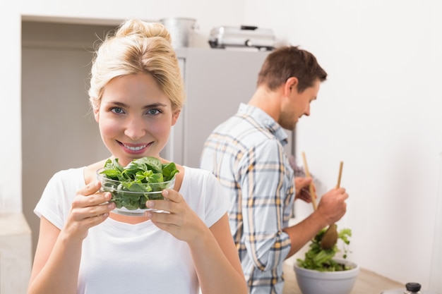 Mulher segurando tigela de folhas com o homem preparando salada no fundo na cozinha