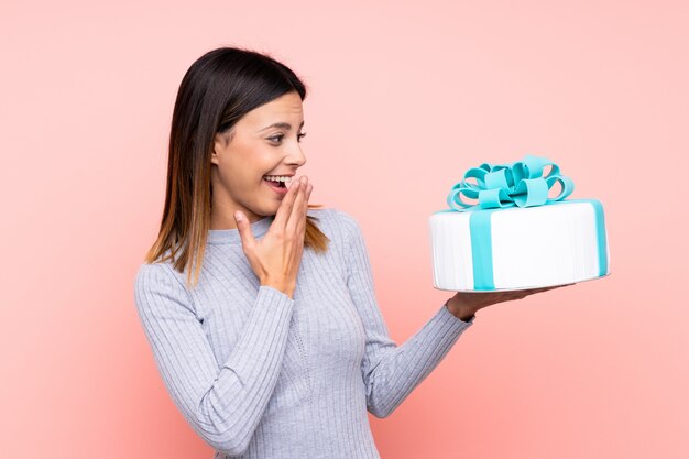 Mulher segurando um bolo grande parede rosa com expressão facial surpreso e chocado