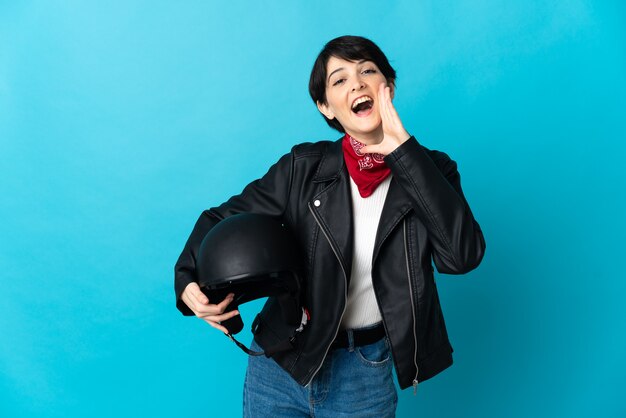 Mulher segurando um capacete de motociclista isolado em um fundo azul, gritando com a boca aberta