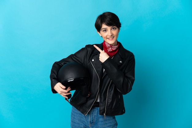 Mulher segurando um capacete de motociclista isolado em uma parede azul apontando para o lado para apresentar um produto