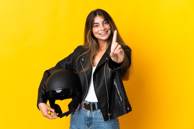 Mulher segurando um capacete de motociclista isolado na parede amarela, mostrando e levantando um dedo