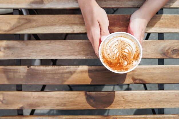 Mulher segurando um copo de papel com café. café para viagem. saborosa bebida quente na mesa de madeira em dia de sol. refeição ao ar livre. vista plana leiga, superior.