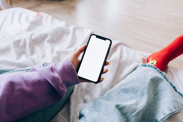 Mulher segurando um smartphone com uma simulação de tela branca, descansando na cama em casa.