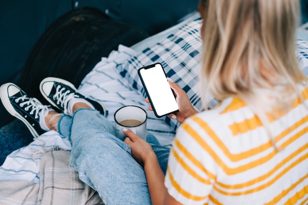 Mulher segurando um smartphone com uma simulação de tela branca, sentado em uma van e bebendo chá durante o intervalo da viagem.