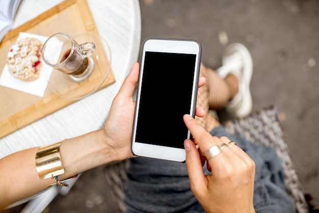 Mulher segurando um telefone inteligente com tela vazia, sentada ao ar livre em um café com bolo e café na mesa