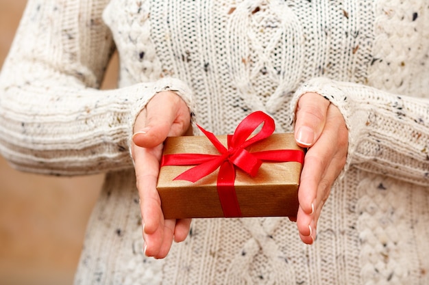 Mulher segurando uma caixa de presente amarrada com uma fita vermelha nas mãos. Profundidade de campo rasa, foco seletivo na caixa. Conceito de dar um presente no feriado ou aniversário.