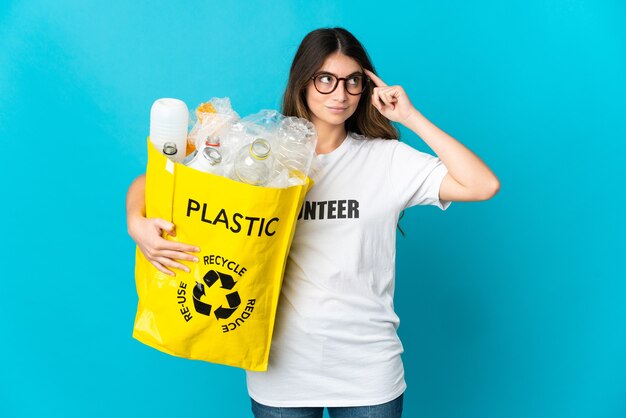 Mulher segurando uma sacola cheia de garrafas para reciclar isolada na parede azul, tendo dúvidas e pensando