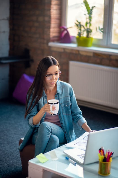 Mulher segurando uma xícara com chá enquanto trabalha no laptop