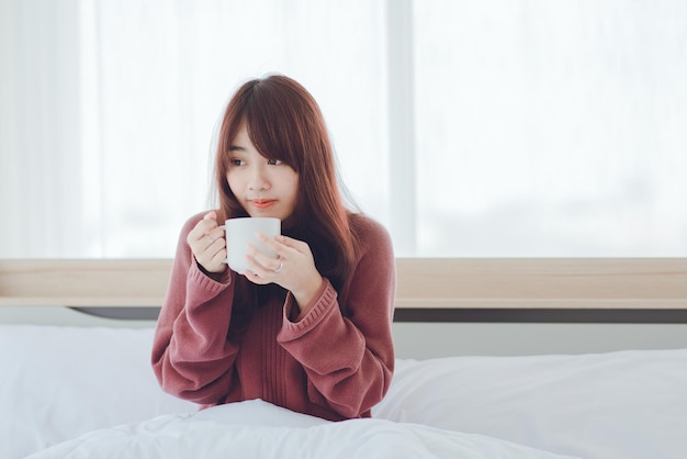 Mulher segurando uma xícara de café com leite na cama em um quarto brancoMulher bebendo