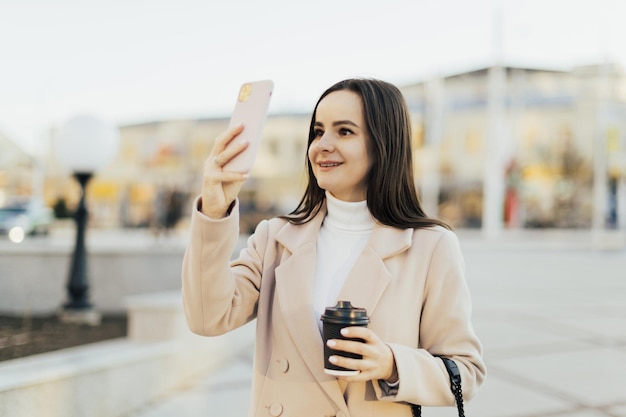 mulher segurando uma xícara quente de café e tirando foto com telefone inteligente na cidade