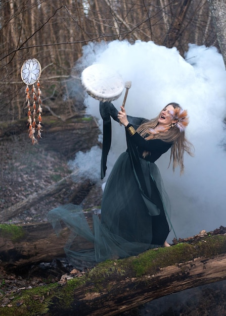 Foto mulher selvagem tocando tambor xamânico de pé no tronco de uma árvore de musgo verde na floresta bomba de fumaça branca