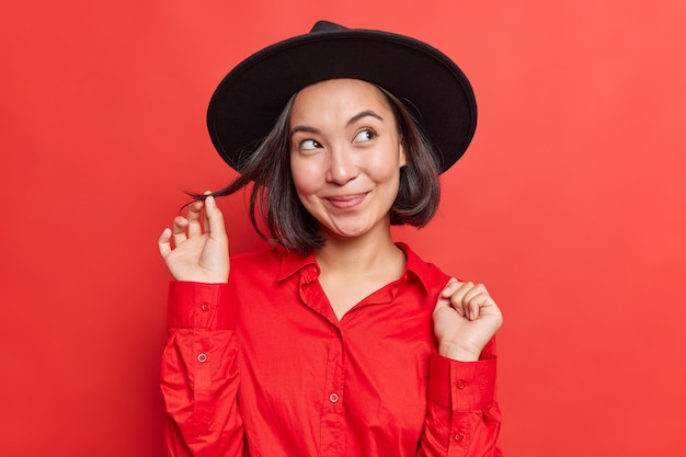 mulher sem maquiagem com expressão sonhadora pensa em algo maravilhoso, cachos, cabelo, chapéu preto, camisa vermelha, posa, interior, vivid studio